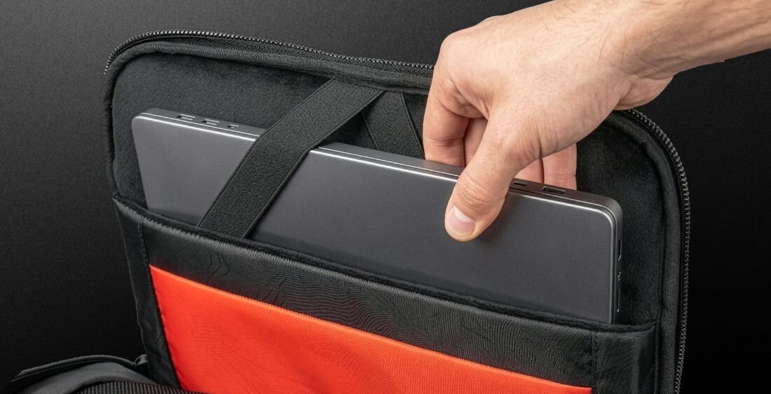 Osoba wkłada laptopa do czarnego plecaka z czerwonym wnętrzem. Plecak posiada elastyczny pasek zabezpieczający laptopa.