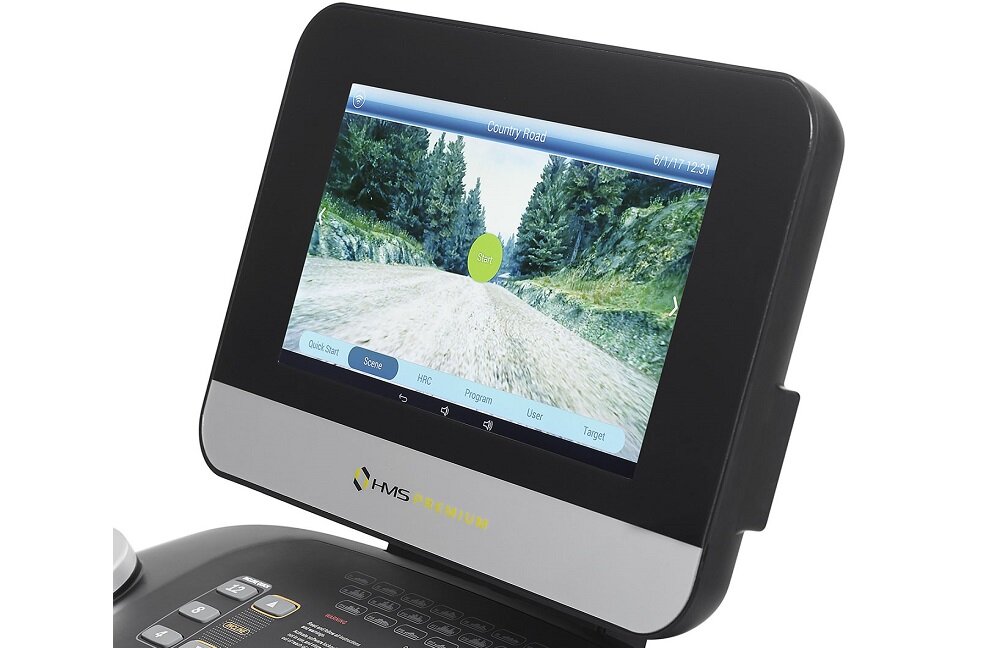 Wyświetlacz bieżni elektrycznej pokazuje symulację biegu po leśnej drodze. Na dole ekranu widoczne są przyciski z napisami: \'Quick Start\', \'Speed\', \'Help\', \'Program\', \'User\', \'Target\'.