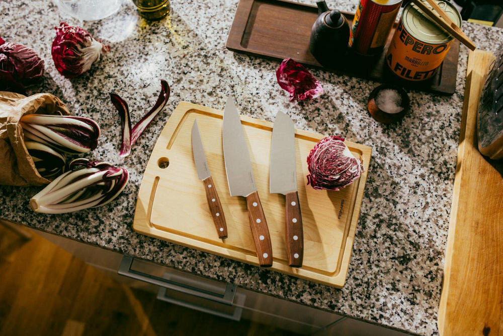 Na blacie kuchennym znajduje się drewniana deska do krojenia z trzema nożami o drewnianych rękojeściach. Obok leżą warzywa, puszka z napisem \'PEPERONI FILETTI\' oraz inne kuchenne akcesoria.