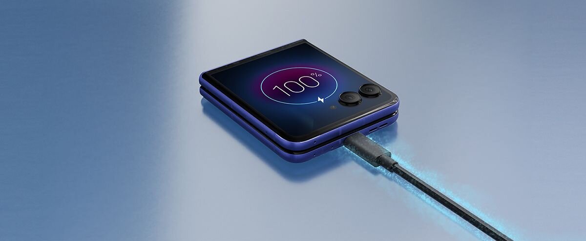 Na obrazie widać złożony smartfon z wyświetlaczem pokazującym 100% naładowania baterii, podłączony do ładowarki. Obok ekranu znajdują się dwa obiektywy aparatu.