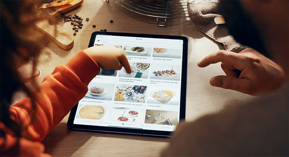 Na stole leży tablet z wyświetlonymi przepisami kulinarnymi, a dwie osoby wskazują na ekran. W tle widoczne są składniki kuchenne i akcesoria.