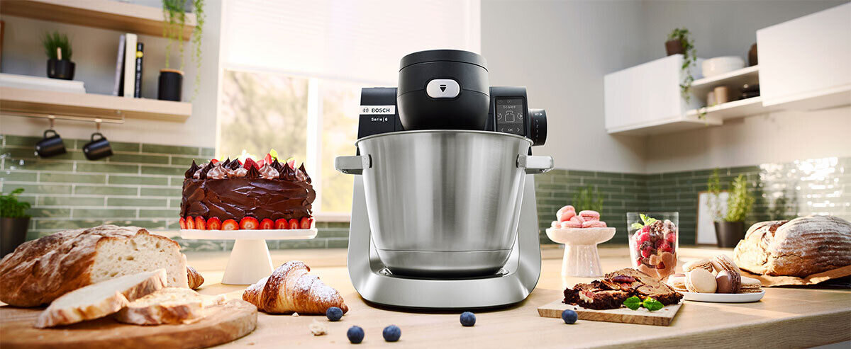 Robot kuchenny planetarny BOSCH z metalową misą stoi na blacie kuchennym, otoczony różnymi wypiekami i owocami. W tle widać jasną kuchnię z zielonymi kafelkami na ścianie.