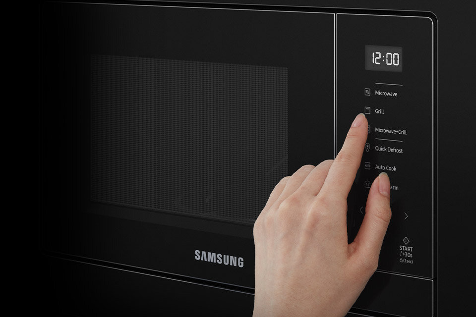 Czarna kuchenka mikrofalowa z panelem dotykowym, na którym widoczne są opcje: Microwave, Grill, Microwave+Grill, Quick Defrost, Auto Cook, Keep Warm. Na wyświetlaczu widnieje godzina 12:00, a poniżej znajduje się logo Samsung.