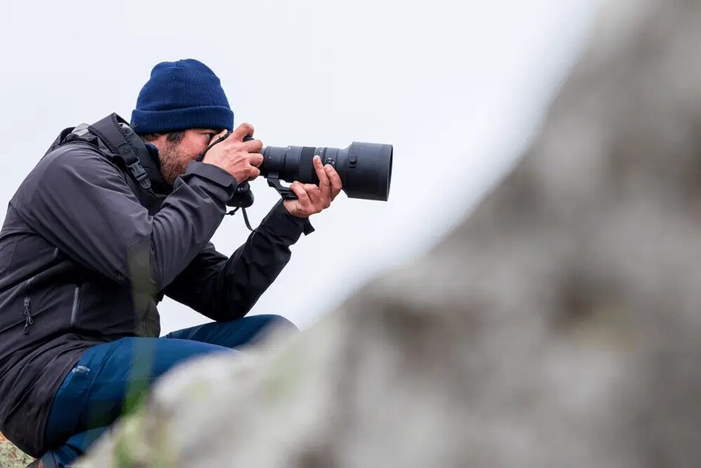 Osoba w ciemnej kurtce i czapce trzyma aparat z dużym obiektywem, siedząc na zewnątrz. W tle widoczna jest rozmyta powierzchnia skały.