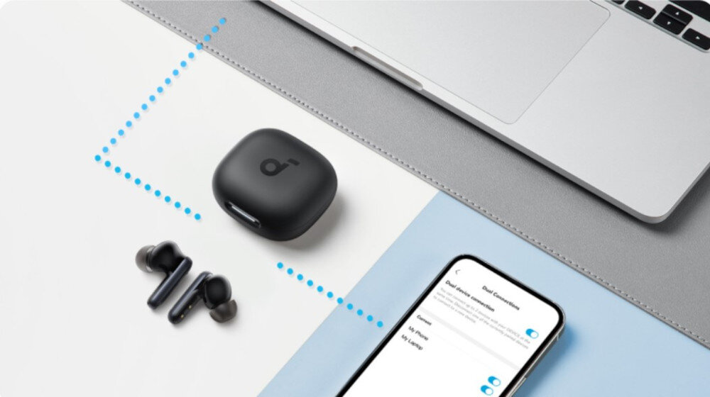 Na obrazie widoczne są czarne słuchawki dokanałowe z etui ładującym, obok znajduje się smartfon z wyświetlonym menu ustawień oraz fragment laptopa. Na etui widnieje logo w kształcie litery \'a\'.