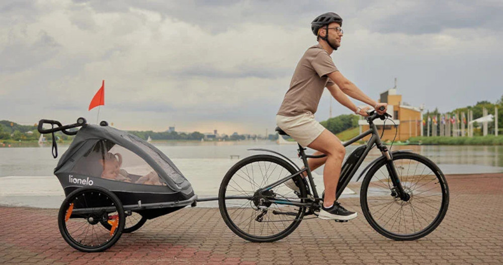 Mężczyzna na rowerze ciągnie przyczepkę rowerową z dzieckiem, na której widnieje napis \'lionelo\'. Przyczepka jest szara z pomarańczowymi elementami i ma czerwoną flagę.