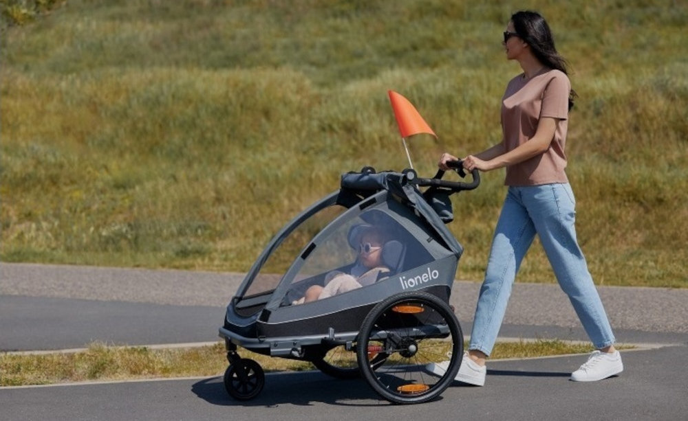 Osoba prowadzi przyczepkę rowerową z dzieckiem w środku, na której widoczne jest logo \'lionelo\'. Przyczepka jest szara z pomarańczową flagą na górze.