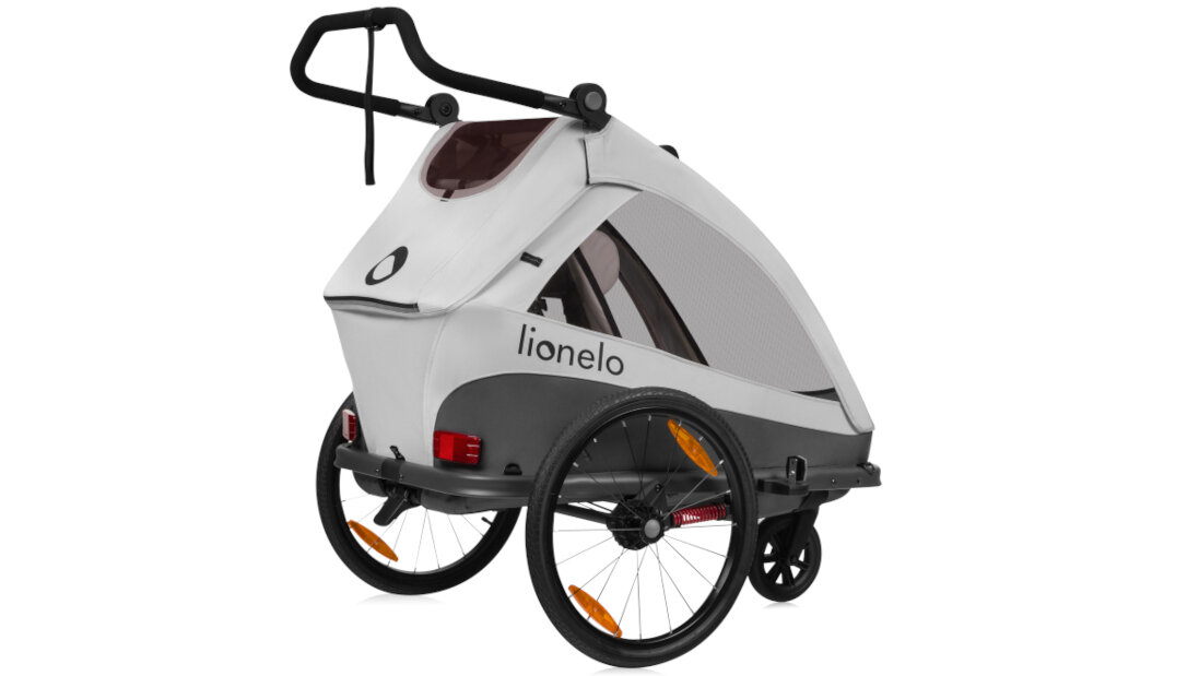 Przyczepka rowerowa w kolorze szarym z czarnymi elementami, posiada dwa koła z pomarańczowymi odblaskami i uchwyt do prowadzenia. Na boku widoczny jest napis \'lionelo\'.