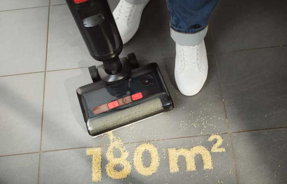 Odkurzacz mopujący zbiera rozsypane drobinki z podłogi, na której widoczny jest napis \'180 m²\'. Obok odkurzacza widać osobę w białych butach stojącą na szarej podłodze.