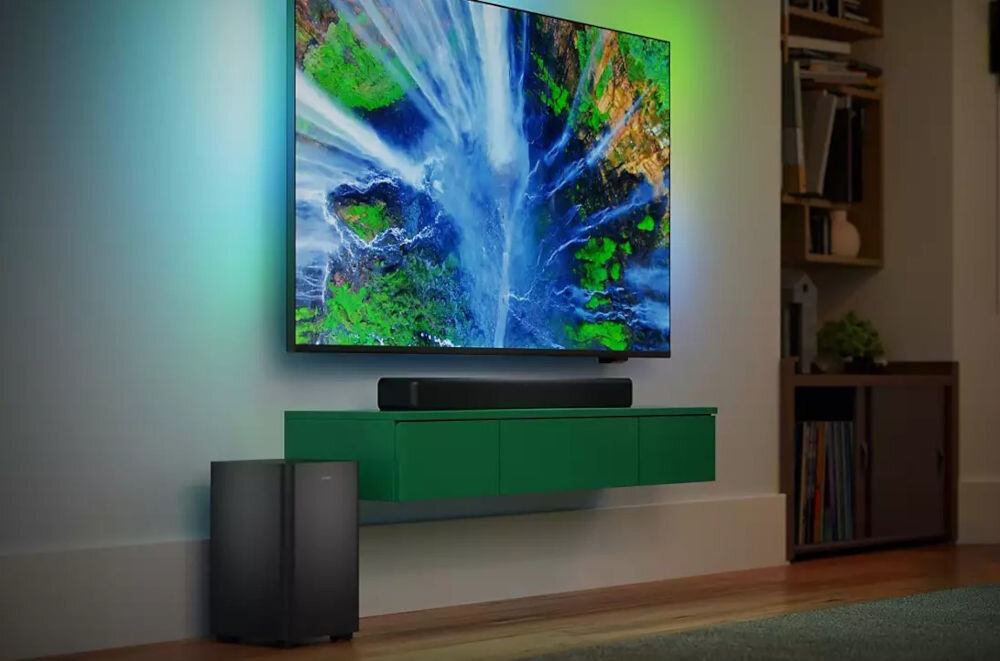 Na obrazie widać nowoczesny salon z dużym telewizorem wyświetlającym kolorowy obraz wodospadu. Pod telewizorem znajduje się soundbar, a obok stoi bezprzewodowy subwoofer.