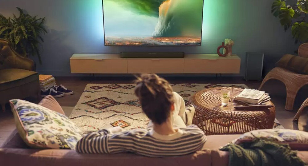 Osoba siedzi na kanapie w salonie, oglądając duży telewizor z dynamiczną sceną przyrody. Na stoliku obok znajduje się szklanka z napojem i książka.