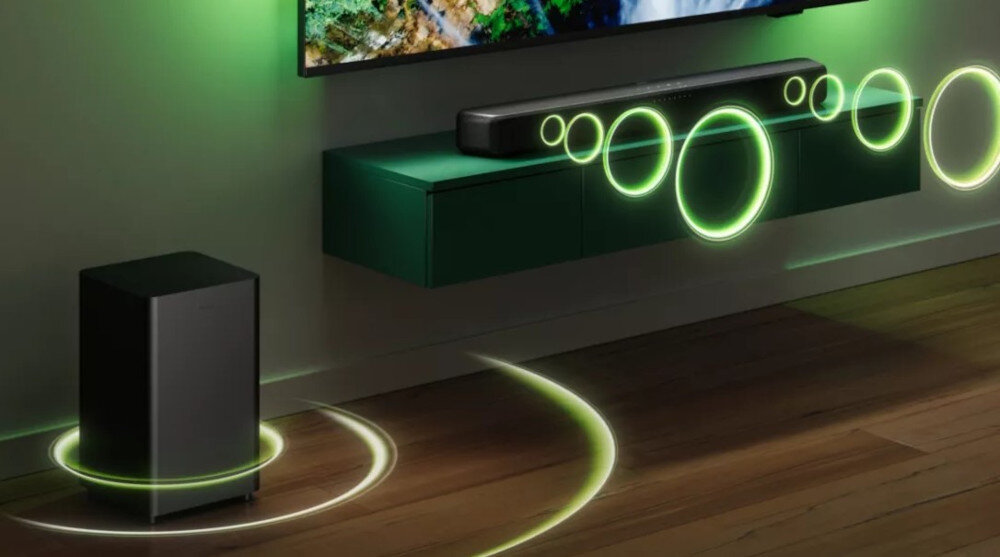Na obrazie widać szary soundbar umieszczony na zielonej półce pod telewizorem oraz czarny subwoofer stojący na podłodze. Wokół urządzeń widoczne są zielone kręgi symbolizujące dźwięk.