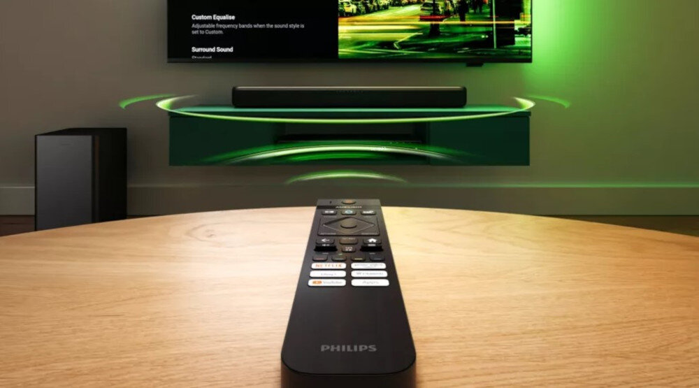 Na drewnianym stole leży pilot z logo Philips, skierowany w stronę telewizora z zielonymi efektami świetlnymi. W tle widoczny jest soundbar i subwoofer, a na ekranie telewizora wyświetlane są opcje dźwięku: \'Custom Equalize\' i \'Surround Sound\'.