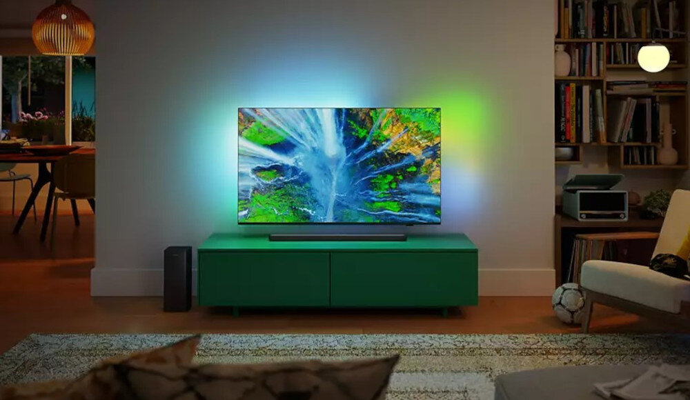 W salonie na zielonej szafce stoi telewizor z włączonym kolorowym obrazem, a przed nim znajduje się soundbar. W tle widać półkę z książkami i lampę.