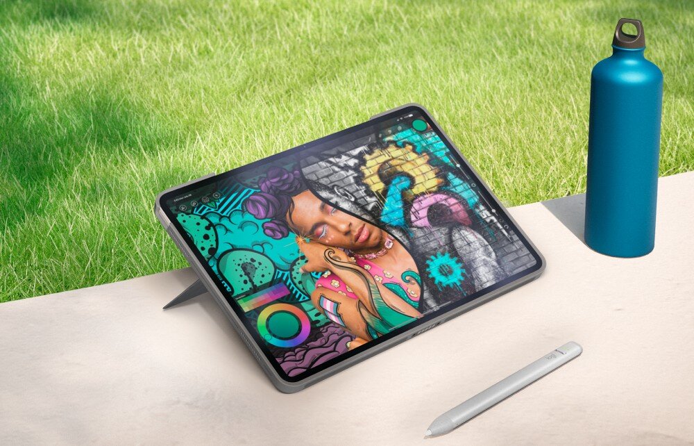 Na stole znajduje się tablet w szarym etui z kolorową grafiką na ekranie, obok leży rysik i niebieska butelka. W tle widoczna jest zielona trawa.
