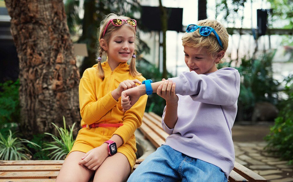 Dwoje dzieci siedzi na ławce w otoczeniu roślin, jedno z nich nosi niebieski smartwatch na nadgarstku. Drugie dziecko wskazuje na zegarek, oboje są uśmiechnięci.