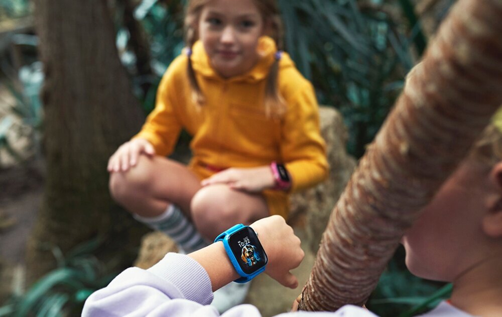Dziecko nosi niebieski smartwatch z kolorowym wyświetlaczem, na którym widoczna jest godzina 10:21. W tle znajduje się inne dziecko w żółtej bluzie.