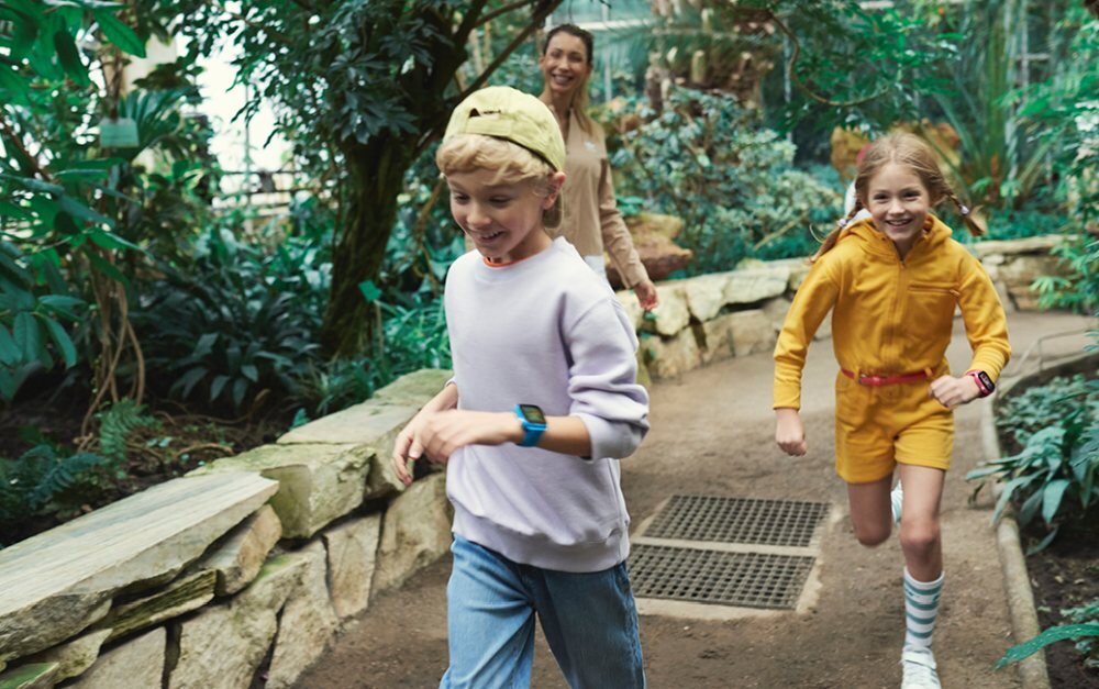Dwoje dzieci biegnie ścieżką w ogrodzie botanicznym, każde z nich ma na nadgarstku smartwatch. W tle widoczna jest osoba dorosła oraz bujna roślinność.