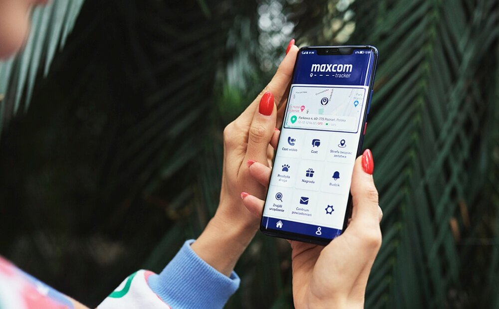 Osoba trzyma smartfon z aplikacją \'maxcom tracker\' na ekranie, na którym widoczne są ikony takie jak \'Czas wideo\', \'Czat\', \'Strefa bezpieczna\' i inne. W tle znajdują się zielone liście roślin.