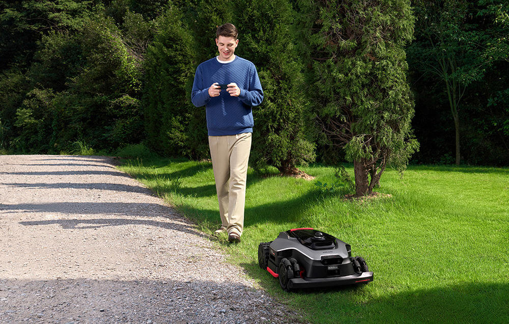 Na trawniku znajduje się czarny robot koszący z czerwonymi elementami. Obok stoi osoba trzymająca urządzenie przypominające pilot.