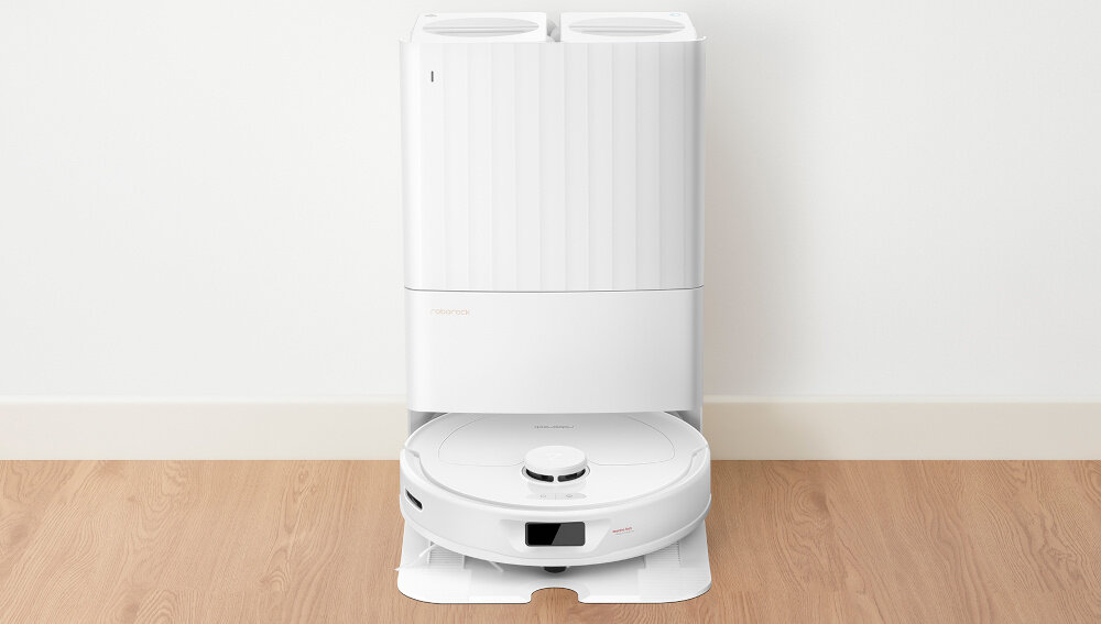 Na drewnianej podłodze stoi biały robot sprzątający z okrągłą stacją dokującą. Na stacji widoczne jest logo \'roborock\'.