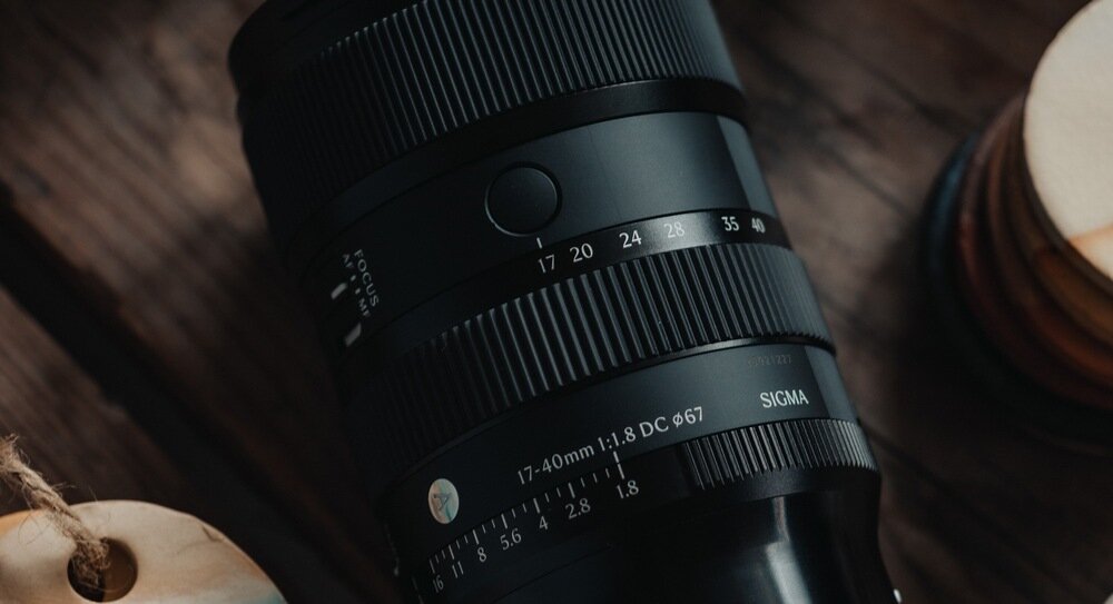 Obiektyw fotograficzny z widocznymi oznaczeniami 17-40mm f/1.8 DC oraz logo SIGMA, leży na drewnianej powierzchni.
