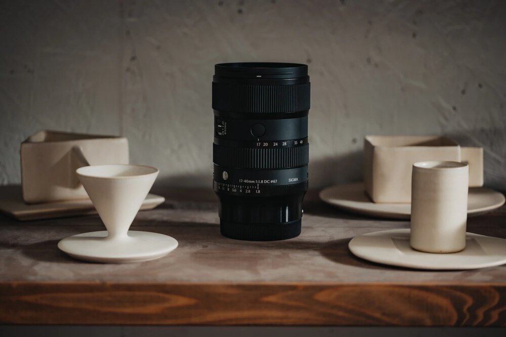 Na drewnianym stole stoi czarny obiektyw z widocznymi oznaczeniami \'17-40mm 1:1.8 DC 82\' oraz \'SIGMA\'. W tle znajdują się ceramiczne naczynia w jasnym kolorze.