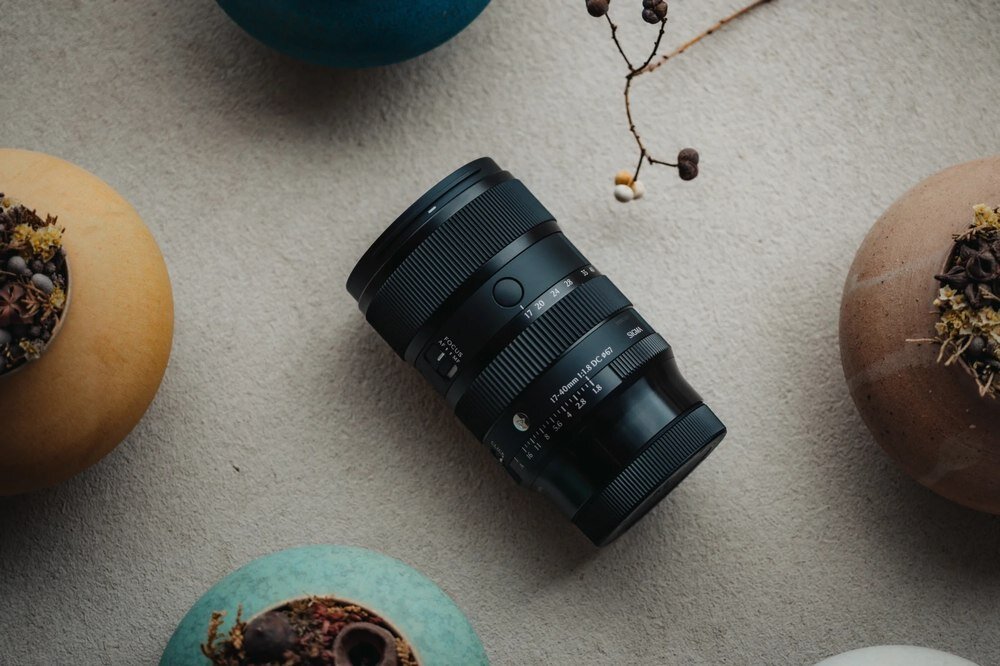 Czarny obiektyw fotograficzny leży na jasnym tle, otoczony trzema ozdobnymi doniczkami z roślinami. Na obiektywie widoczny jest napis: \'17-40mm 1:1.8 DC\'.
