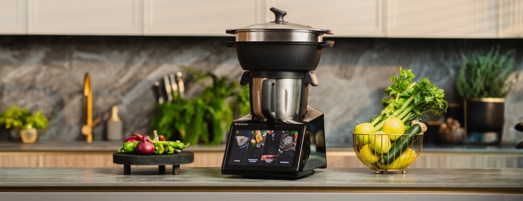 Na blacie kuchennym stoi wielofunkcyjny robot kuchenny z wyświetlaczem, obok którego znajdują się świeże warzywa i owoce. W tle widoczna jest stylowa kuchnia z roślinami ozdobnymi.