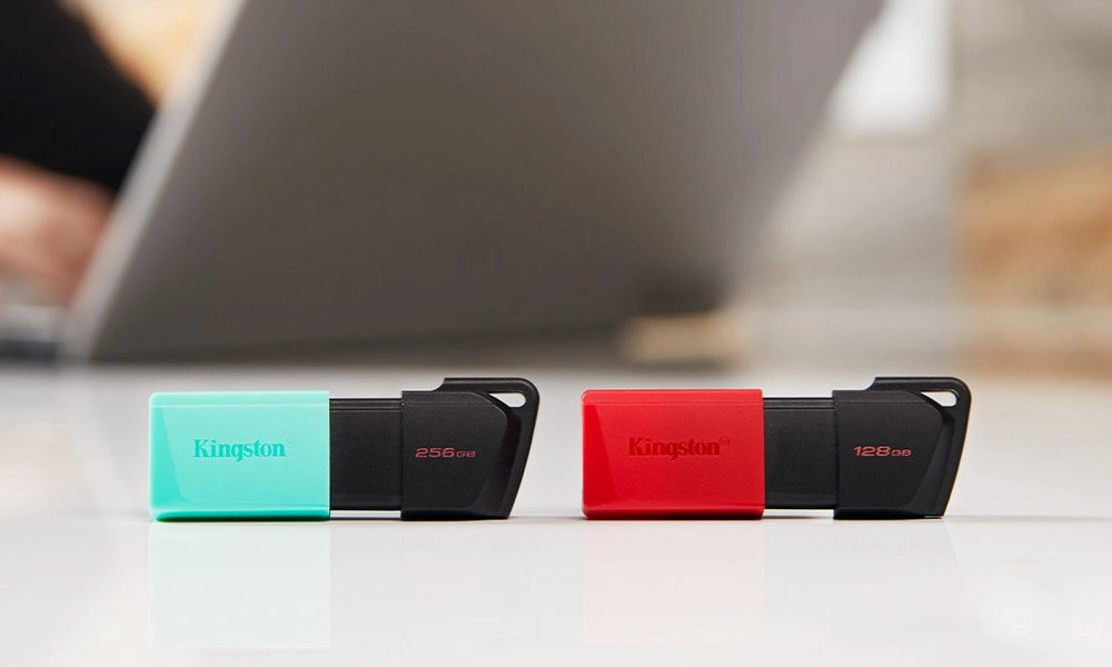 Na obrazie znajdują się dwa pendrive\'y marki Kingston, jeden czarno-turkusowy z napisem \'256GB\', a drugi czarno-czerwony z napisem \'128GB\'. W tle widoczny jest rozmyty laptop.