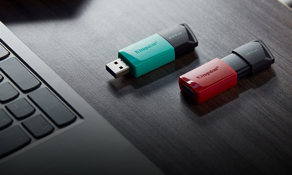 Na drewnianym blacie leżą dwa pendrive\'y, jeden w kolorze czarno-turkusowym z napisem \'Kingston\', a drugi w kolorze czarno-czerwonym. Obok znajduje się fragment klawiatury laptopa.