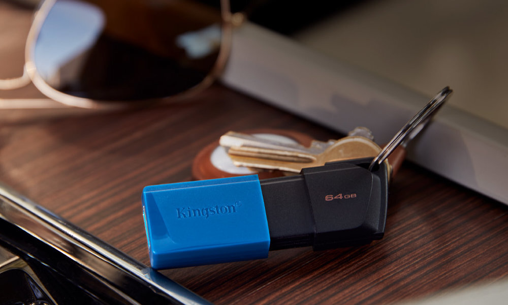 Na drewnianej powierzchni leży czarno-turkusowy pendrive z napisem \'Kingston\' oraz klucze. Obok znajduje się para okularów przeciwsłonecznych.