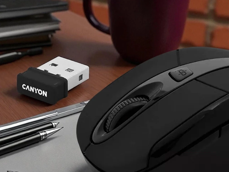 Na drewnianym biurku znajduje się czarna mysz bezprzewodowa z widocznym przyciskiem \'dpi\' oraz odbiornik USB z logo \'CANYON\'. Obok leżą długopisy i kubek.