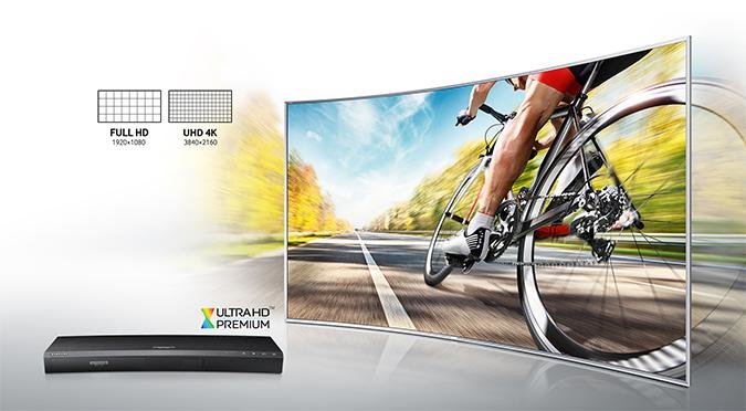 Zakrzywiony ekran telewizora SUHD, na którym wyświetlany jest doskonałej jakości obraz jadącego kolarza. W lewym dolnym rogu widoczny odtwarzacz UHD Blu-ray K8500. W lewym górnym rogu grafika porównująca rozdzielczość Full HD i UHD 4K.