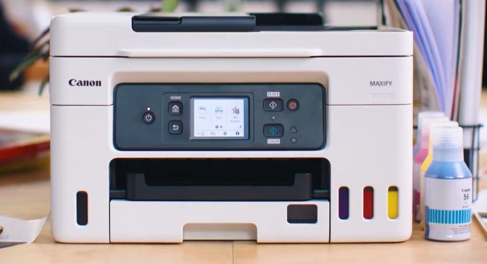 Na obrazie widoczne jest urządzenie wielofunkcyjne marki Canon z wyświetlaczem i przyciskami na panelu przednim. Obok urządzenia znajdują się butelki z tuszem w różnych kolorach.