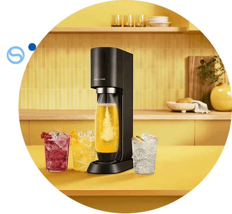 Czarny saturator z logo \'sodastream\' stoi na żółtym blacie, obok trzech szklanek z kolorowymi napojami. W tle znajduje się żółta ściana i półka z naczyniami.