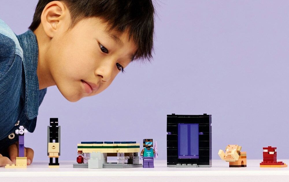 Na białym stole znajdują się klocki LEGO przedstawiające różne postacie i konstrukcje, w tym portal z fioletowym wnętrzem. W tle widać dziecko wpatrujące się w klocki na fioletowym tle.
