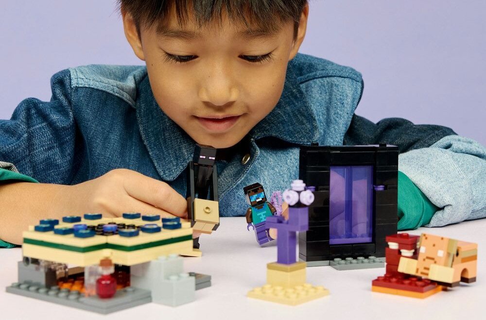 Dziecko bawi się zestawem klocków LEGO, przedstawiającym sceny z gry Minecraft. Widoczne są różne konstrukcje i postacie z klocków, w tym portal i figurki postaci.