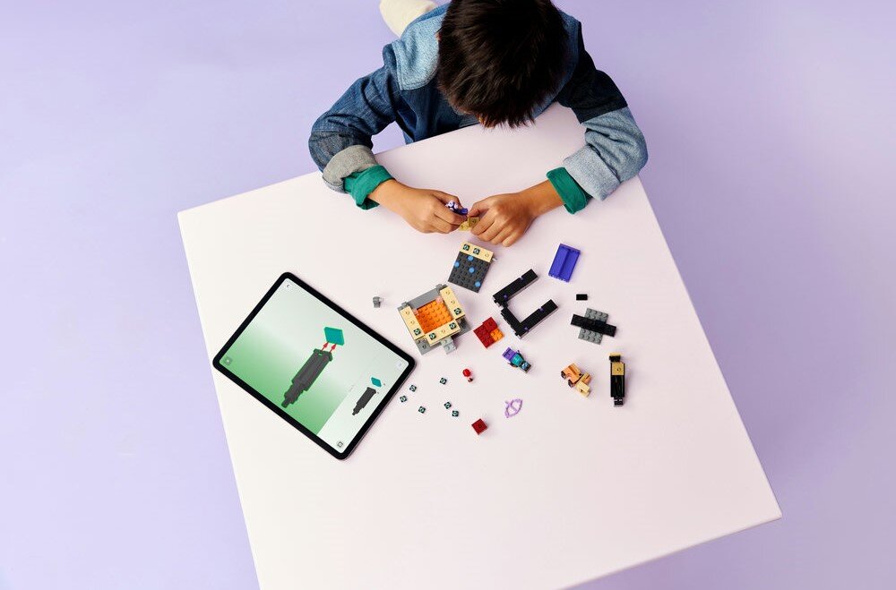 Dziecko siedzi przy białym stole, na którym znajdują się klocki LEGO oraz tablet z wyświetlonym obrazem. Klocki są różnokolorowe i ułożone w różne kształty.