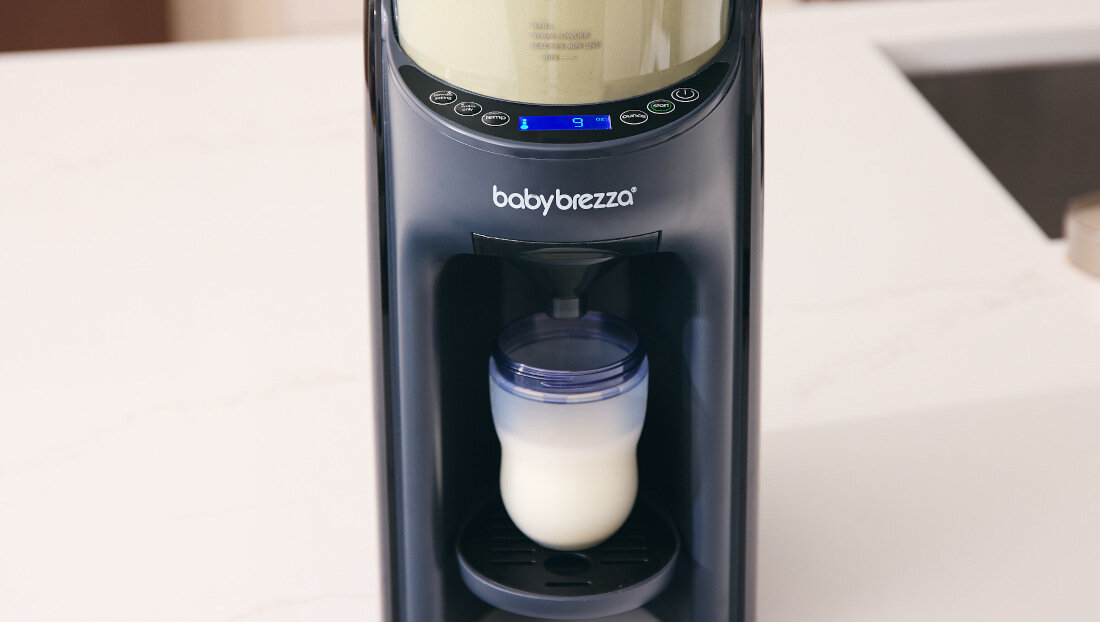 Na blacie kuchennym stoi ekspres do mleka z logo \'babybrezza\', w którym znajduje się butelka z mlekiem. Na panelu widoczny jest wyświetlacz z cyfrą \'9\'.