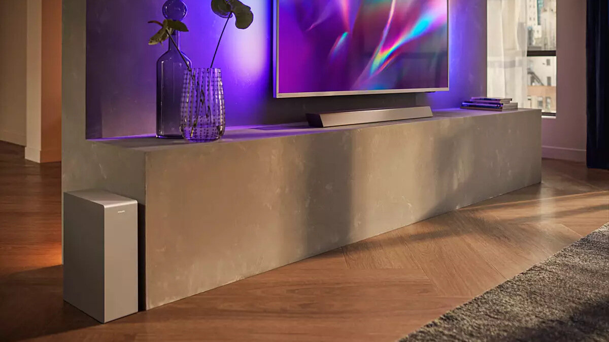 Na obrazie widać nowoczesny salon z telewizorem na szafce, pod którym znajduje się srebrny soundbar. Obok szafki stoi subwoofer, a na szafce są dekoracyjne wazony i książki.