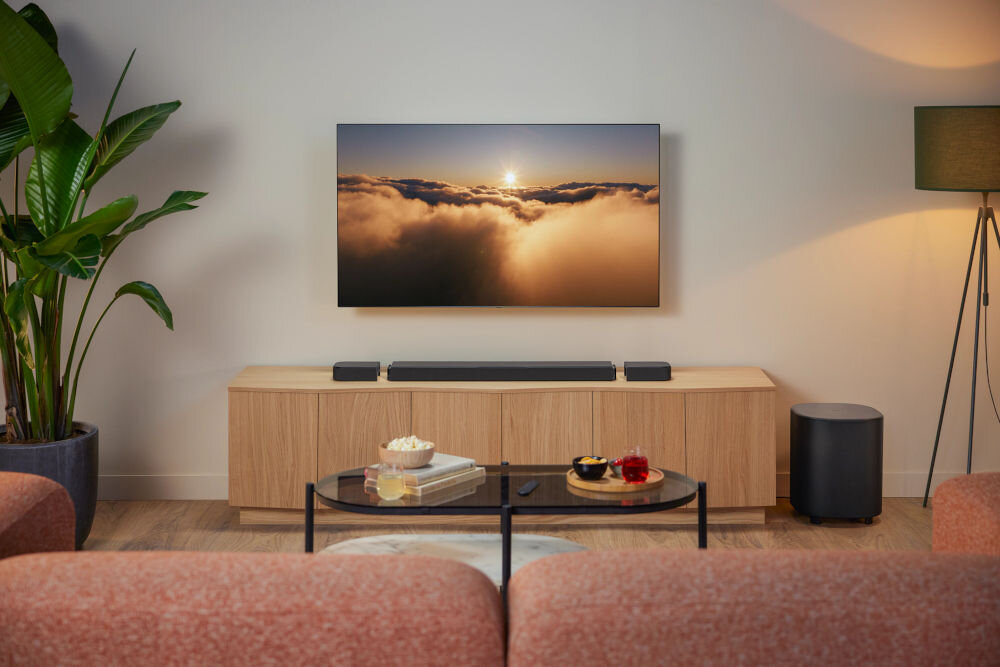 W salonie znajduje się telewizor wyświetlający obraz chmur o zachodzie słońca, umieszczony na ścianie nad drewnianą szafką. Na szafce stoi soundbar, a obok znajduje się bezprzewodowy subwoofer; w pomieszczeniu są także rośliny i lampa stojąca.