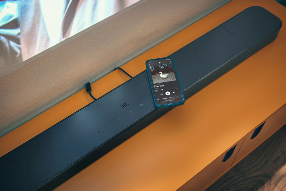Na pomarańczowej powierzchni znajduje się czarny soundbar z logo JBL. Na soundbarze leży smartfon z wyświetlonym ekranem odtwarzacza muzyki.
