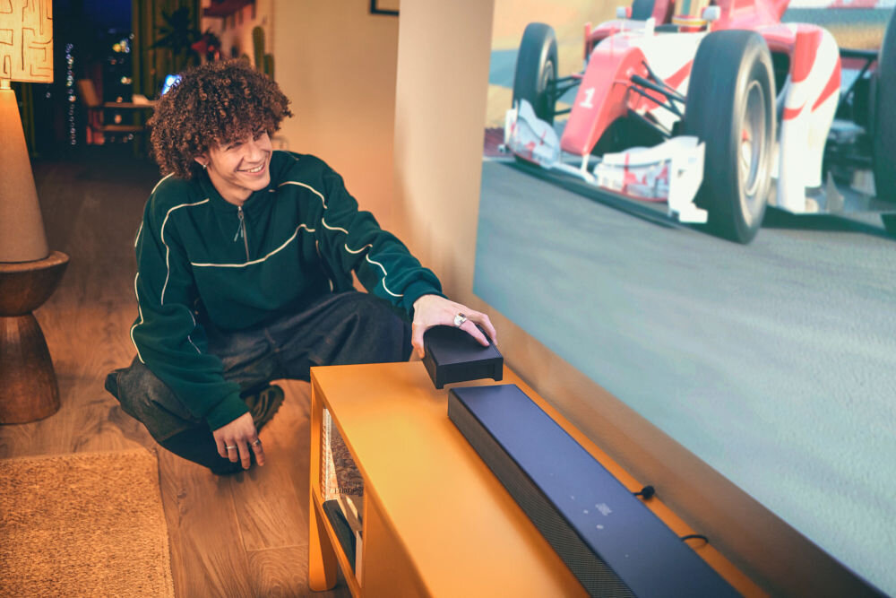 Osoba siedzi na podłodze obok stolika, na którym znajduje się czarny soundbar i mały głośnik. W tle widać ekran z wyścigowym samochodem Formuły 1.
