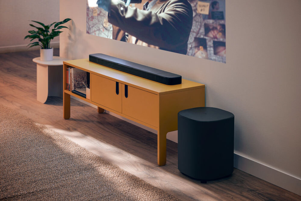 Na zdjęciu znajduje się czarny soundbar umieszczony na żółtej szafce, obok której stoi czarny subwoofer. W tle widać fragment projekcji filmowej na ścianie oraz roślinę na białym stoliku.