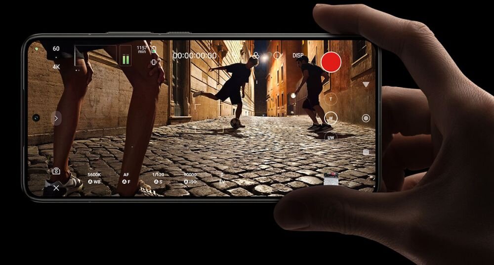 Osoba trzyma smartfon, na ekranie którego widać nagranie wideo przedstawiające dwie osoby grające w piłkę na brukowanej ulicy. Na ekranie widoczne są różne ikony i wskaźniki związane z nagrywaniem wideo.