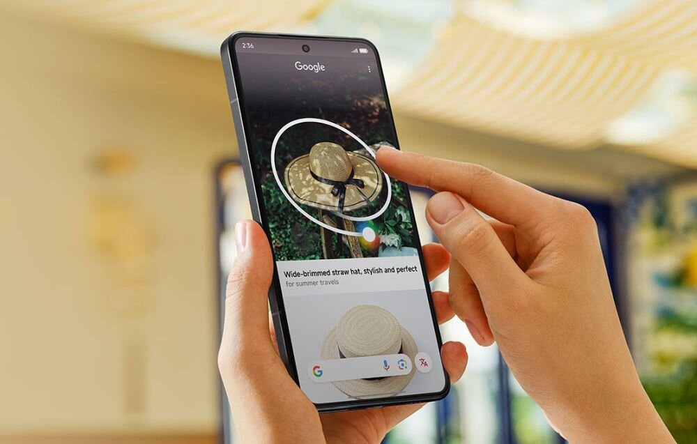 Osoba trzyma smartfon, na ekranie którego widać wyszukiwarkę Google z obrazem kapelusza. Pod zdjęciem znajduje się tekst: \'Wide-brimmed straw hat, stylish and perfect for summer travels\'.