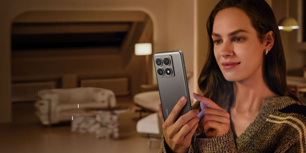 Osoba trzyma smartfon z czterema kamerami z tyłu, w tle widać nowoczesne wnętrze z sofą i lampą.