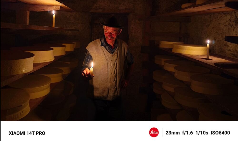 Mężczyzna trzyma świecę w ciemnym pomieszczeniu z półkami pełnymi okrągłych serów. Na dole obrazu widoczny jest tekst: \'XIAOMI 14T PRO\', logo \'Leica\' oraz parametry: \'23mm f/1.6 1/10s ISO6400\'.