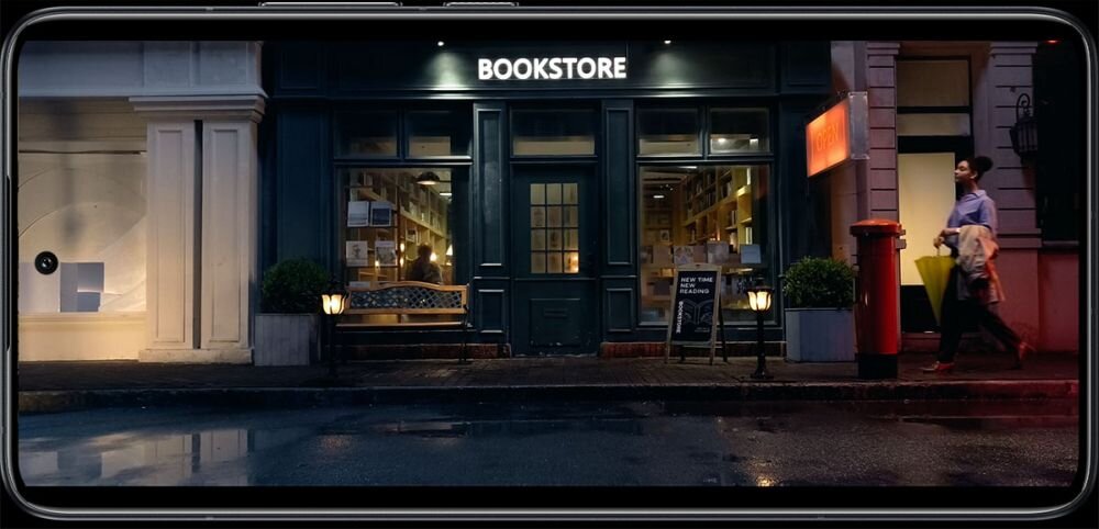 Na ekranie smartfona widać fasadę księgarni z napisem \'BOOKSTORE\'. Przed wejściem stoi ławka, a obok przechodzi osoba z torbą na zakupy.